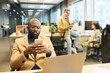 © pressmaster - Serious African American manager using smartphone while sitting by workplace in front of laptop and texting against two colleagues