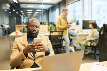 Fototapete - Serious African American manager using smartphone while sitting by workplace in front of laptop and texting against two colleagues