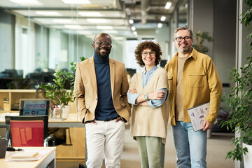 Poster - Three successful managers or brokers in smart casualwear looking at camera while standing in openspace office with green plants