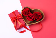 Three Red Rosebuds In A Wooden Bowl In The Shape Of A Heart With Red Gift Box On A Pink And Red Background. Happy Valentine's Day, Mother's Day And Birthday Greeting Card.
