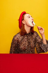 Wall Mural - Beautiful young girl in stylish red beret tasting whipped cream over yellow studio background. Food pop art photography. Complementary colors. Concept of art, beauty, food. Copy space for ad, text.