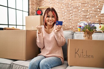 Wall Mural - Young caucasian woman moving to a new home holding credit card screaming proud, celebrating victory and success very excited with raised arms