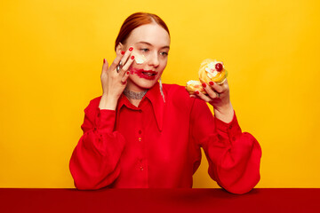 Wall Mural - Surrealism. Beautiful young girl in red shirt with smudged red lipstick, eating cake over yellow studio background. Food pop art photography. Complementary colors. Concept of art, beauty, food.