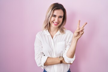 Poster - Young beautiful woman standing over pink background smiling with happy face winking at the camera doing victory sign with fingers. number two.