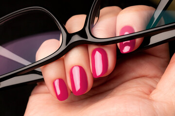 Wall Mural - Female hand with beautiful manicure - viva magenta, pink nails with glasses on black background closeup