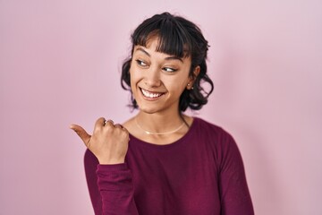 Sticker - Young beautiful woman standing over pink background smiling with happy face looking and pointing to the side with thumb up.