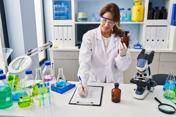 Canvas Print - Young latin woman wearing scientist uniform writing on clipboard at laboratory