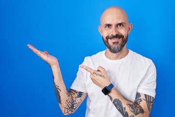 Poster - Hispanic man with tattoos standing over blue background amazed and smiling to the camera while presenting with hand and pointing with finger.