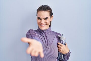 Poster - Beautiful woman wearing sportswear holding water bottle smiling cheerful offering palm hand giving assistance and acceptance.