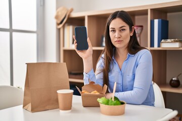 Sticker - Young brunette woman eating take away food at home showing smartphone screen thinking attitude and sober expression looking self confident