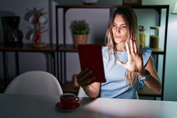 Sticker - Young hispanic woman using touchpad sitting on the table at night doing stop sing with palm of the hand. warning expression with negative and serious gesture on the face.