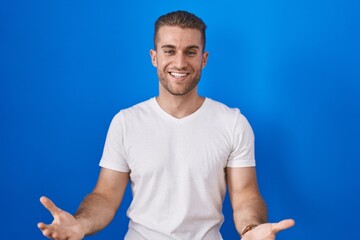 Sticker - Young caucasian man standing over blue background smiling cheerful with open arms as friendly welcome, positive and confident greetings