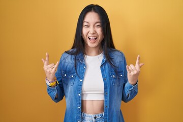 Canvas Print - Young asian woman standing over yellow background shouting with crazy expression doing rock symbol with hands up. music star. heavy music concept.