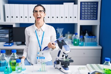 Canvas Print - Young brunette woman working at scientist laboratory angry and mad screaming frustrated and furious, shouting with anger. rage and aggressive concept.