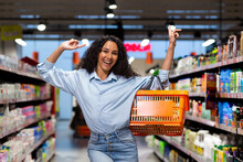 Portrait Of A Happy And Smiling Woman Shopper In A Supermarket, Herpanic Woman Shopping And Looking At The Camera Happily Chooses Products Inside The Store Among The Shelves With A Shopping Basket.