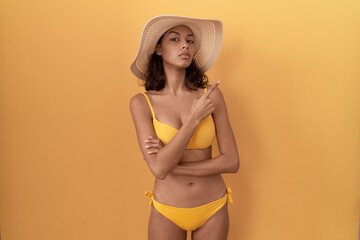 Canvas Print - Young hispanic woman wearing bikini and summer hat pointing with hand finger to the side showing advertisement, serious and calm face