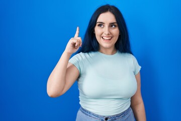 Wall Mural - Young modern girl with blue hair standing over blue background pointing finger up with successful idea. exited and happy. number one.