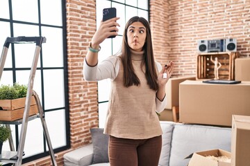 Sticker - Young brunette woman moving to a new home doing video call making fish face with mouth and squinting eyes, crazy and comical.