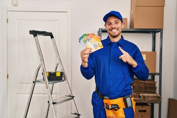 Poster - Young hispanic man working at renovation holding swiss banknotes smiling happy pointing with hand and finger to the side