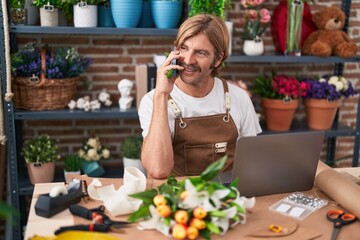 Canvas Print - Young blond man florist talking on smartphone using laptop at flower shop