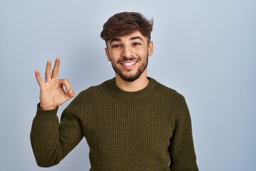 Poster - Arab man with beard standing over blue background smiling positive doing ok sign with hand and fingers. successful expression.