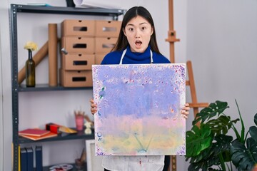 Wall Mural - Chinese young woman holding canvas at art studio afraid and shocked with surprise and amazed expression, fear and excited face.