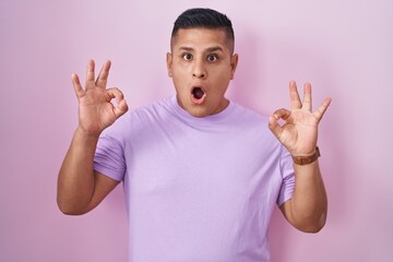 Canvas Print - Young hispanic man standing over pink background looking surprised and shocked doing ok approval symbol with fingers. crazy expression