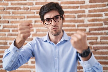 Canvas Print - Young hispanic man standing over brick wall background angry and mad raising fists frustrated and furious while shouting with anger. rage and aggressive concept.