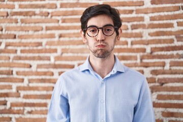 Poster - Young hispanic man standing over brick wall background puffing cheeks with funny face. mouth inflated with air, crazy expression.