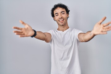 Wall Mural - Hispanic man standing over isolated background looking at the camera smiling with open arms for hug. cheerful expression embracing happiness.