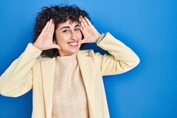 Sticker - Young brunette woman with curly hair standing over blue background smiling cheerful playing peek a boo with hands showing face. surprised and exited