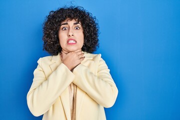 Poster - Young brunette woman with curly hair standing over blue background shouting suffocate because painful strangle. health problem. asphyxiate and suicide concept.