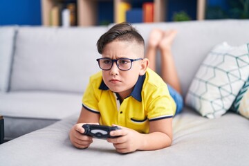 Wall Mural - Young hispanic kid playing video game holding controller on the sofa skeptic and nervous, frowning upset because of problem. negative person.