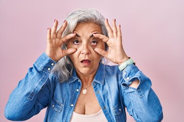 Wall Mural - Middle age woman with grey hair standing over pink background trying to open eyes with fingers, sleepy and tired for morning fatigue