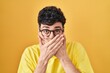 Hispanic man wearing glasses standing over yellow background shocked covering mouth with hands for mistake. secret concept.