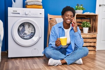 Poster - African american woman talking on the smartphone and drinking coffee waiting for washing machine at laundry room