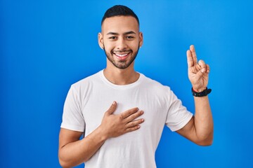 Canvas Print - Young hispanic man standing over blue background smiling swearing with hand on chest and fingers up, making a loyalty promise oath