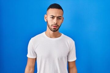 Wall Mural - Young hispanic man standing over blue background looking sleepy and tired, exhausted for fatigue and hangover, lazy eyes in the morning.