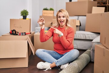 Canvas Print - Blonde woman holding keys of new home sitting on the floor smiling happy pointing with hand and finger