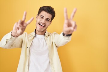 Canvas Print - Young hispanic man standing over yellow background smiling with tongue out showing fingers of both hands doing victory sign. number two.