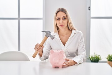 Wall Mural - Young blonde woman holding piggy bank and hammer skeptic and nervous, frowning upset because of problem. negative person.