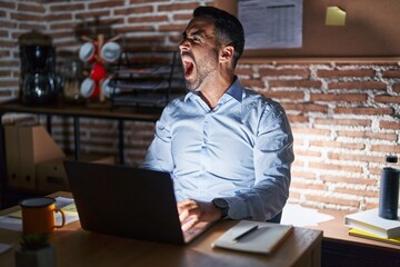 Wall Mural - Hispanic man with beard working at the office at night angry and mad screaming frustrated and furious, shouting with anger. rage and aggressive concept.