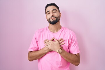 Canvas Print - Hispanic young man standing over pink background smiling with hands on chest with closed eyes and grateful gesture on face. health concept.