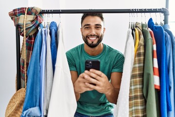 Sticker - Young arab man customer using smartphone appearing through clothes rack at clothing store