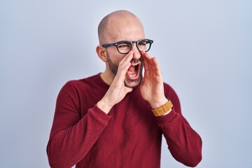 Wall Mural - Young bald man with beard standing over white background wearing glasses shouting angry out loud with hands over mouth