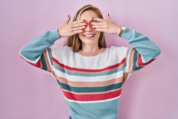 Canvas Print - Young blonde woman standing over pink background covering eyes with hands smiling cheerful and funny. blind concept.