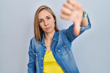 Poster - Young blonde woman standing over blue background looking unhappy and angry showing rejection and negative with thumbs down gesture. bad expression.