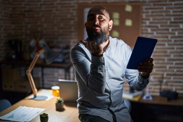 Canvas Print - Young hispanic man with beard and tattoos working at the office at night looking at the camera blowing a kiss with hand on air being lovely and sexy. love expression.