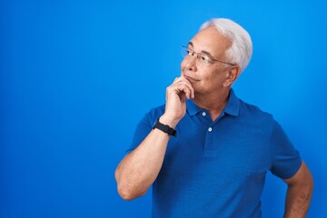 Canvas Print - Middle age man with grey hair standing over blue background with hand on chin thinking about question, pensive expression. smiling with thoughtful face. doubt concept.