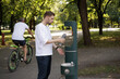 © Girts - A man in a white shirt refills his water bottle at a public refill station in a sustainable city. The station provides tap water to reduce plastic bottle waste. Green city and tap water